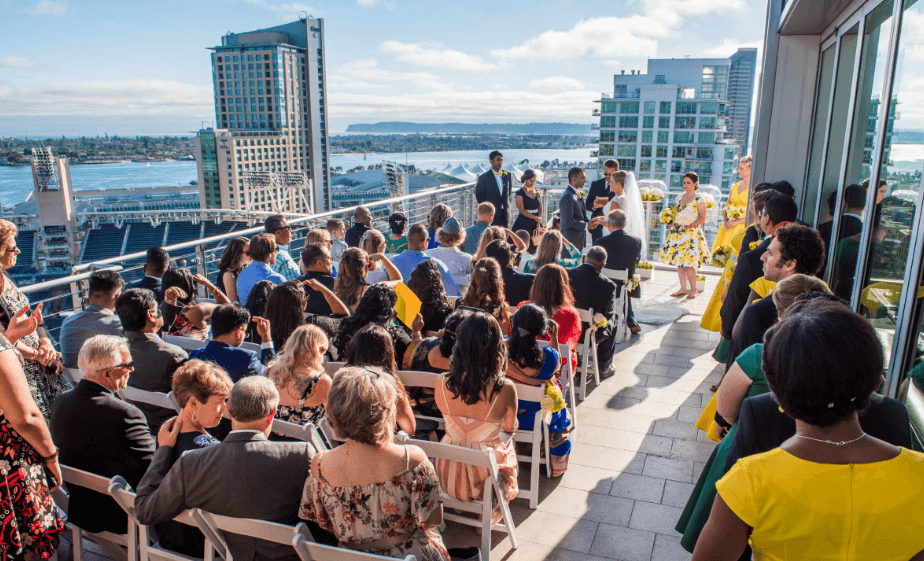 Ceremony on a San Diego rooftop, with guests attentively watching a couple exchange vows, while the city's skyline and waterscape serve as a picturesque backdrop